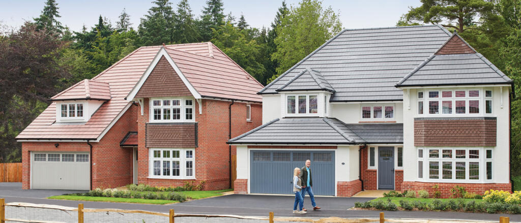 Two people walking in front of two adjacent modern houses with red and grey tiled roofs, surrounded