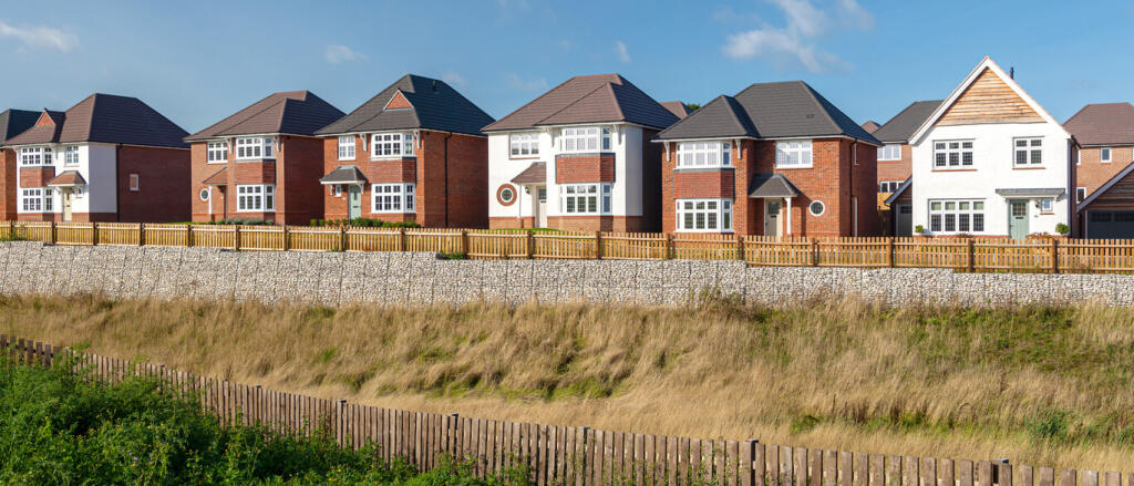 Modern suburban houses with brick and white exteriors, set on a hill behind a wooden fence and a gra