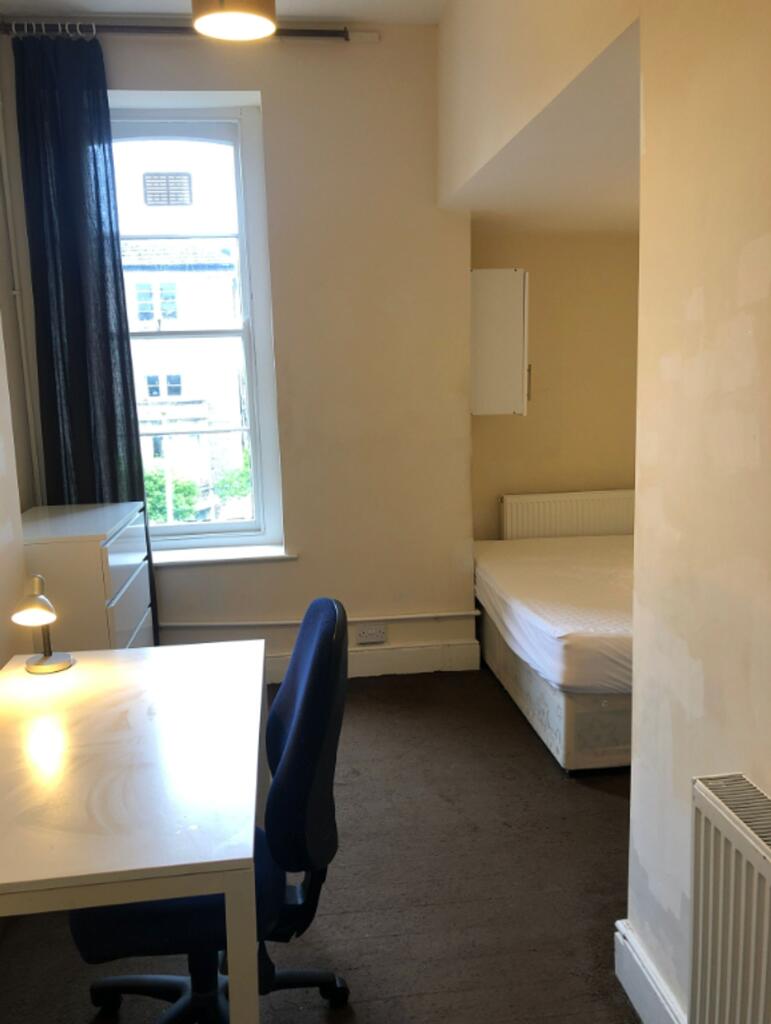 A well-lit single bedroom featuring a desk and ...